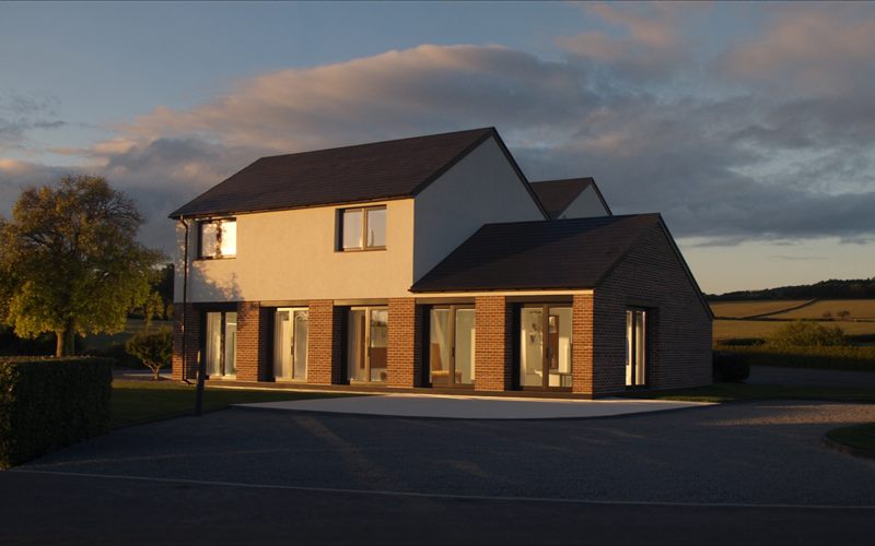 A contemporary UK new-build house with a brick ground floor and white rendered upper storey, lit by low golden-hour sun with open farmland behind