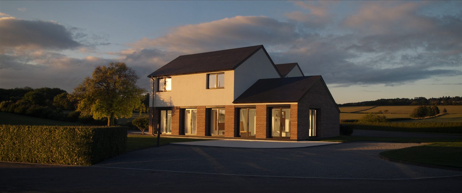 A contemporary UK new-build house with a brick ground floor and white rendered upper storey, lit by low golden-hour sun with open farmland behind