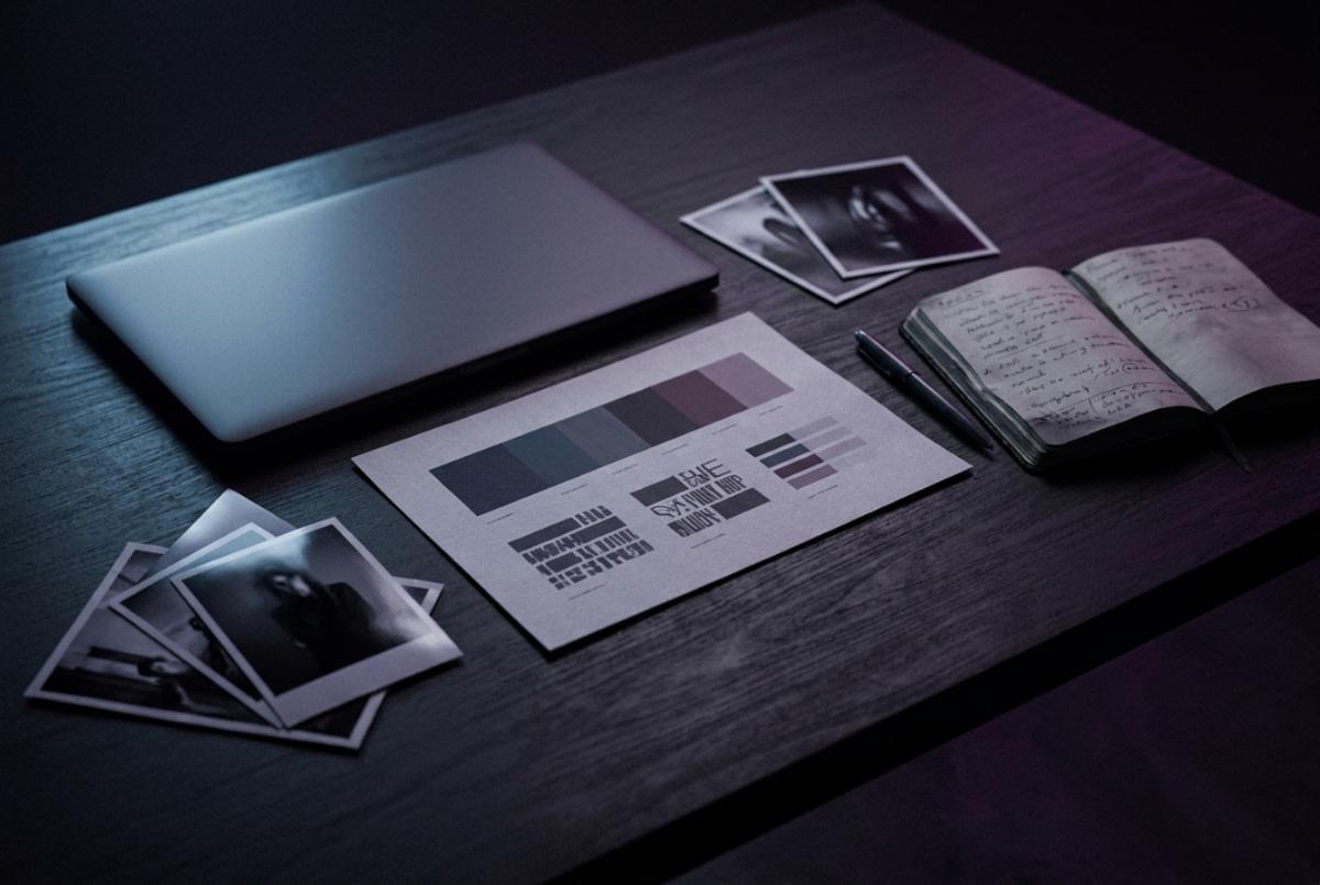 Brand assets laid out on a dark desk: a closed laptop, a printed colour and type specimen sheet, photo prints and an open notebook of project notes