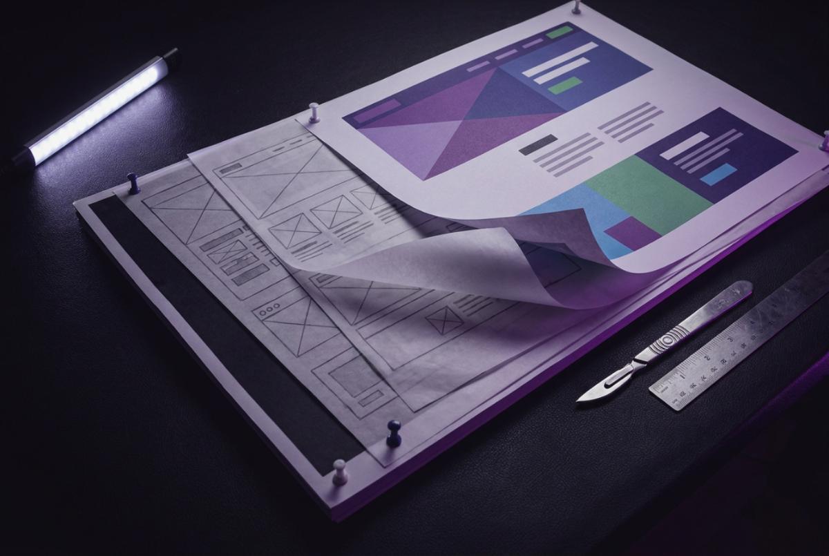 Printed website design boards on a dark surface, a pencil wireframe sheet overlaying a finished colour layout