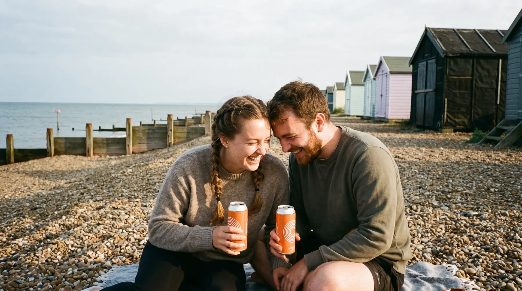 Two friends laughing on a shingle beach with cans of Mardle, beach huts behind them