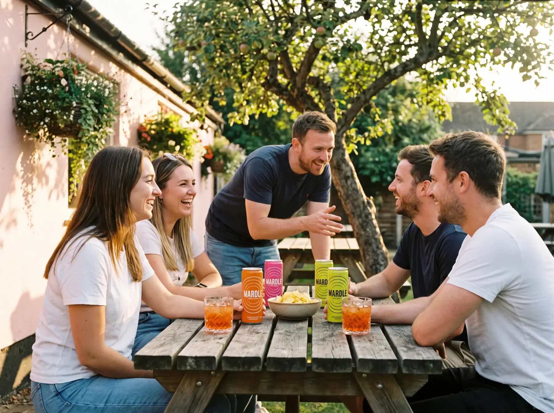 Five friends round a picnic table in a Suffolk pub garden at golden hour, cans of Mardle and tumblers of iced cocktail between them, one leaning in telling a story